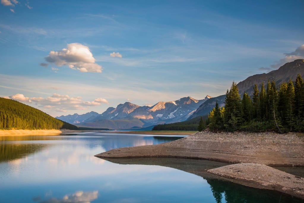 Ruhiger Bergsee mit Nadelwald und spiegelnden Gipfeln im warmen Abendlicht in Kananaskis Country, Kanada.