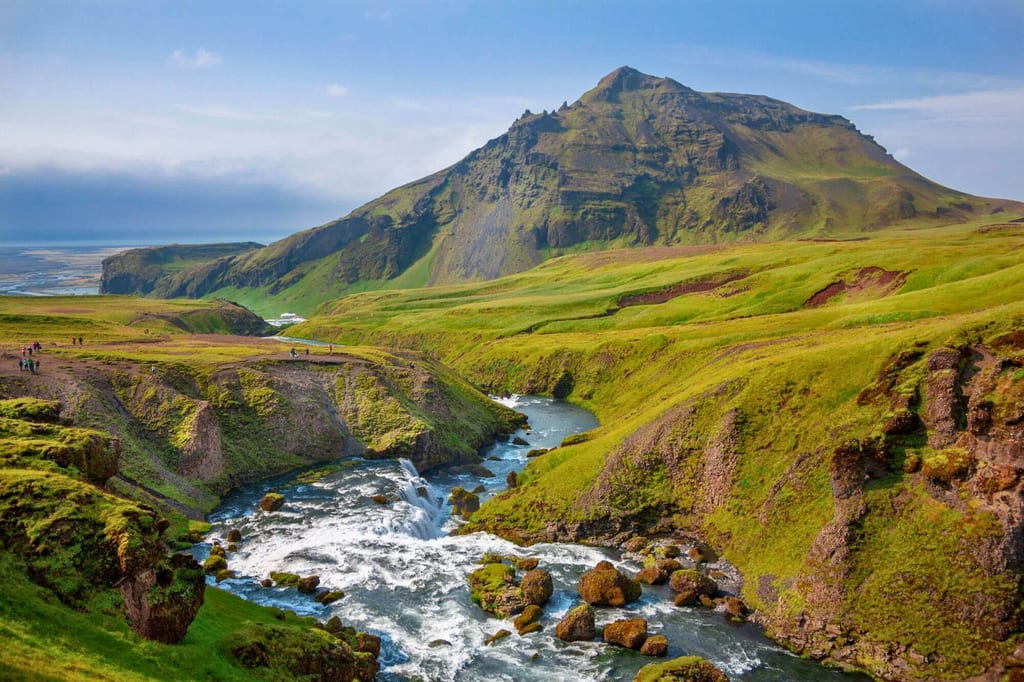 Weite grüne Hügellandschaft mit Fluss, kleinen Wasserfällen und markantem Berg im Hintergrund