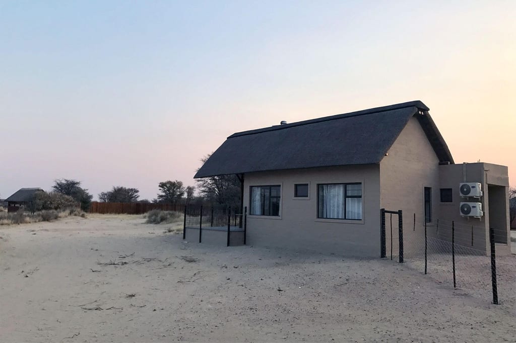 Reetdachhaus auf sandigem Gelände bei ruhigem Abendlicht, Zaun und Klimageräte sichtbar.