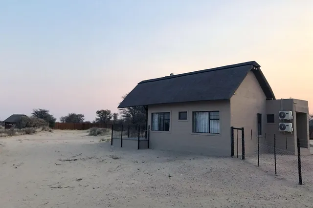 Reetdachhaus auf sandigem Gelände bei ruhigem Abendlicht, Zaun und Klimageräte sichtbar.