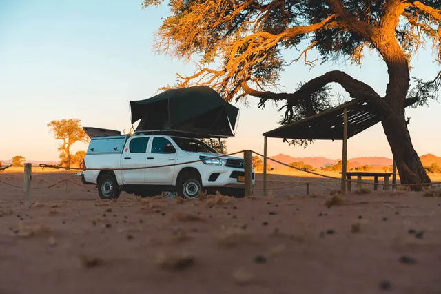 Weißer Geländewagen mit offenem Dachzelt unter einem Baum in sandiger Wüstenlandschaft bei goldenem Licht.