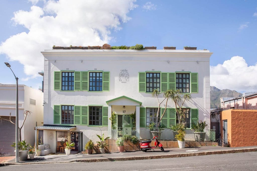 Weiße Hausfassade mit grünen Läden, Topfpflanzen und rotem Motorroller bei sonnigem Himmel.