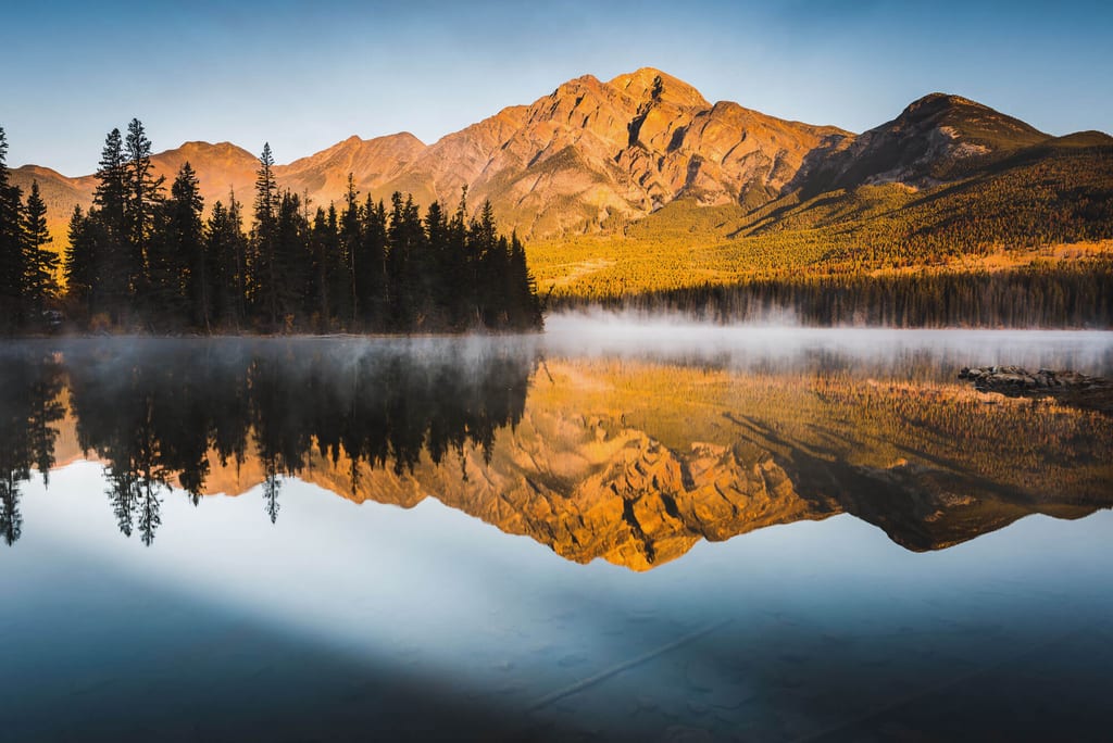 Bergpanorama mit Nadelwald und Spiegelung im See bei Morgennebel und goldenem Licht.