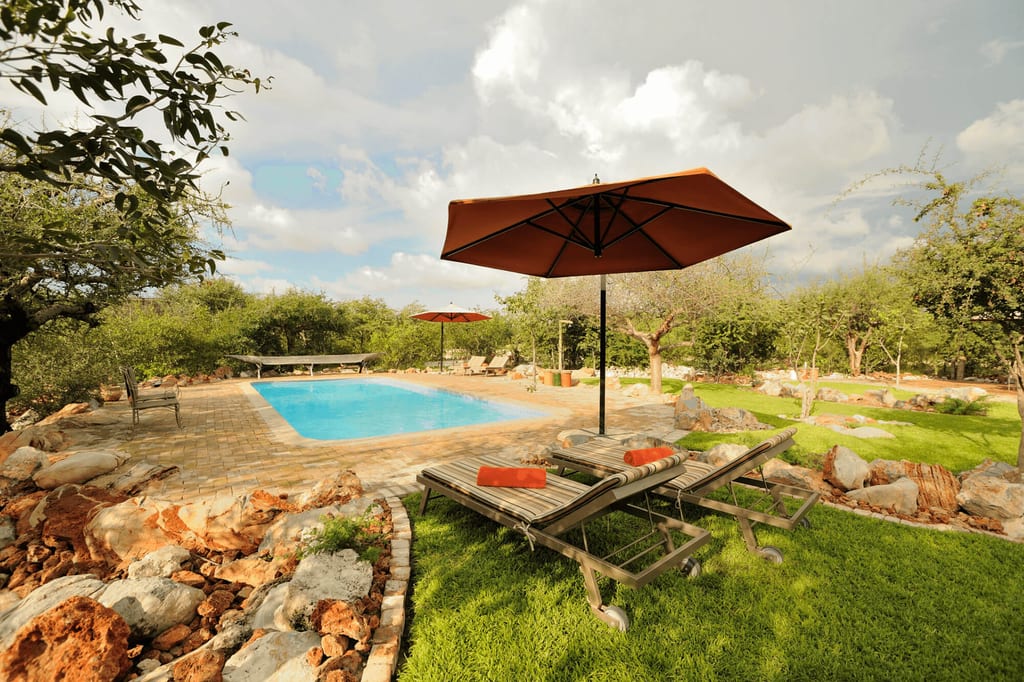 Rechteckiger Pool mit Liegen, Sonnenschirm, Rasen und Felsbeeten unter wechselndem Himmel.