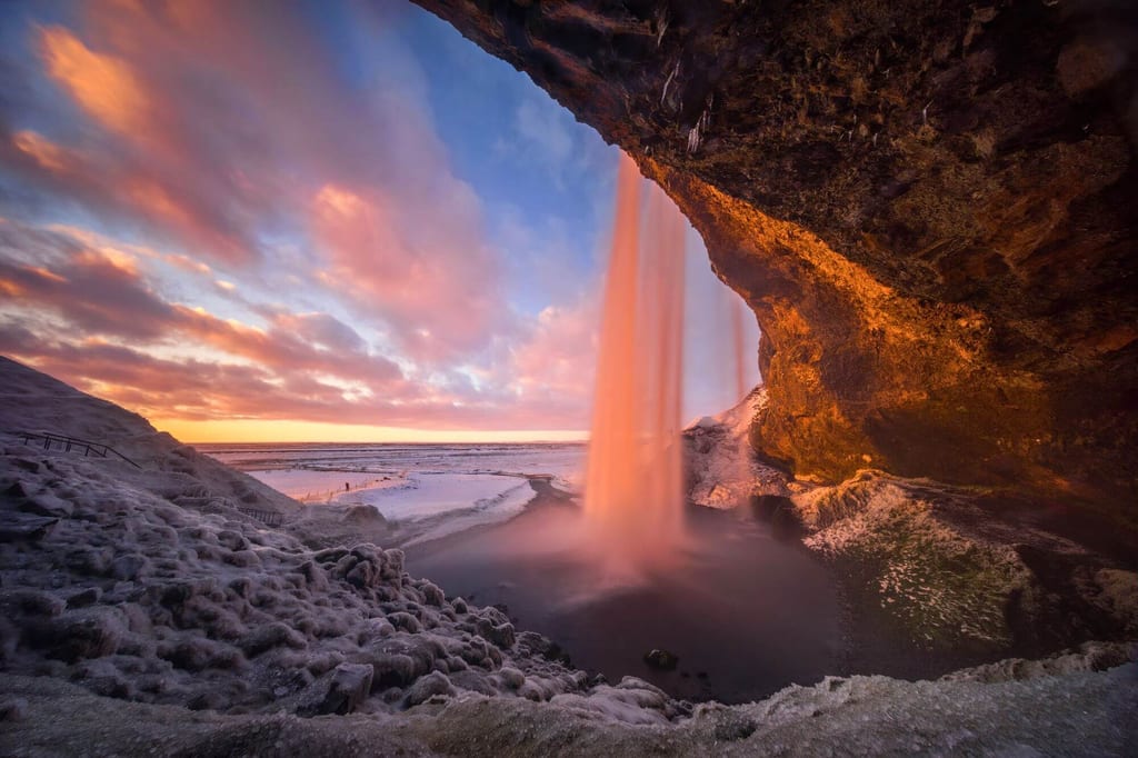 Wasserfall stürzt vor einer eisigen Küstenlandschaft, beleuchtet vom warmen Licht des Sonnenuntergangs.