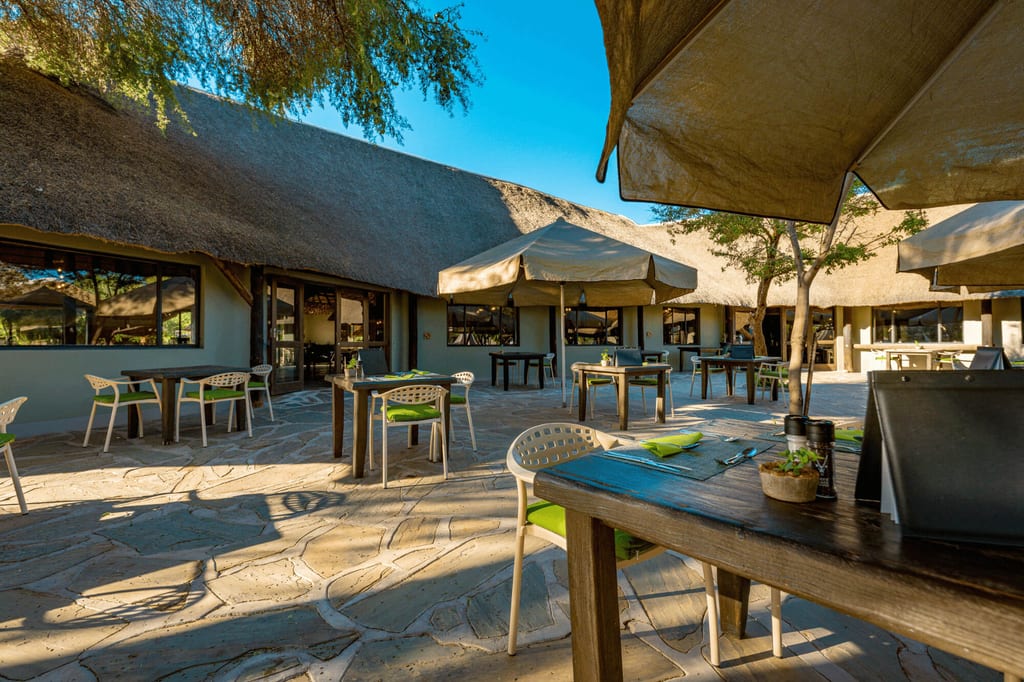 Freiluft-Terrasse einer Lodge mit Strohdach, Tischen und Sonnenschirmen im warmen Morgenlicht.