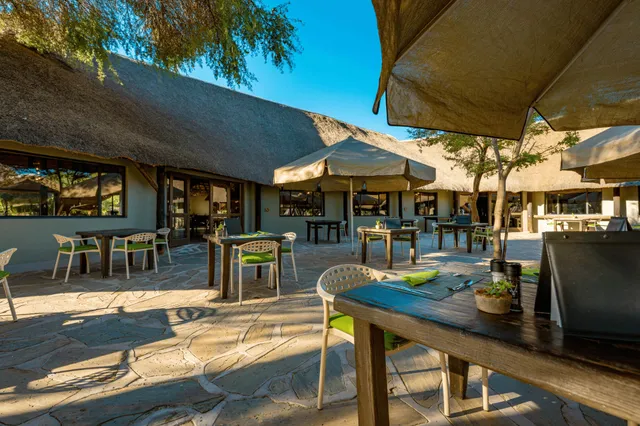 Freiluft-Terrasse einer Lodge mit Strohdach, Tischen und Sonnenschirmen im warmen Morgenlicht.