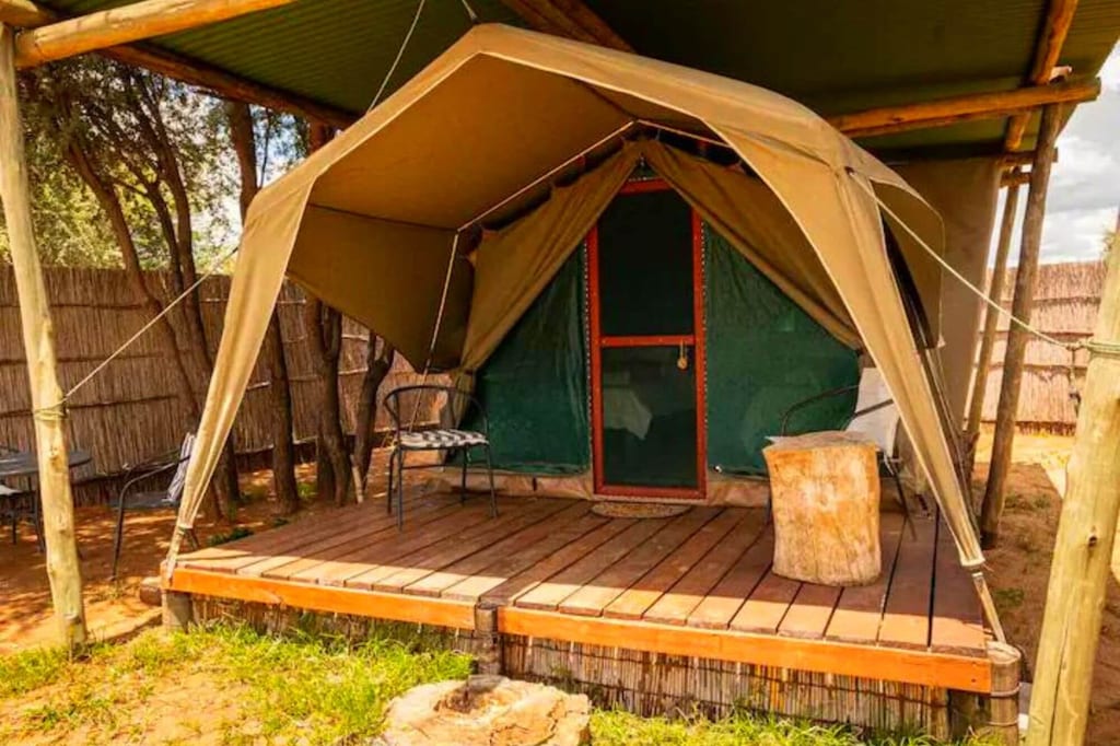 Safari-Zelt auf Holzveranda mit Stühlen, Baumstumpf und Schilfzaun bei sonnigem Tageslicht.