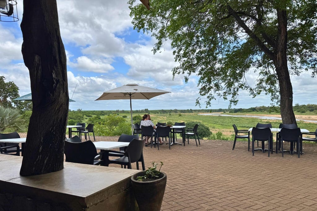 Terrasse mit Tischen und Stühlen, zwei Personen unter Sonnenschirm, Blick auf grüne Flusslandschaft