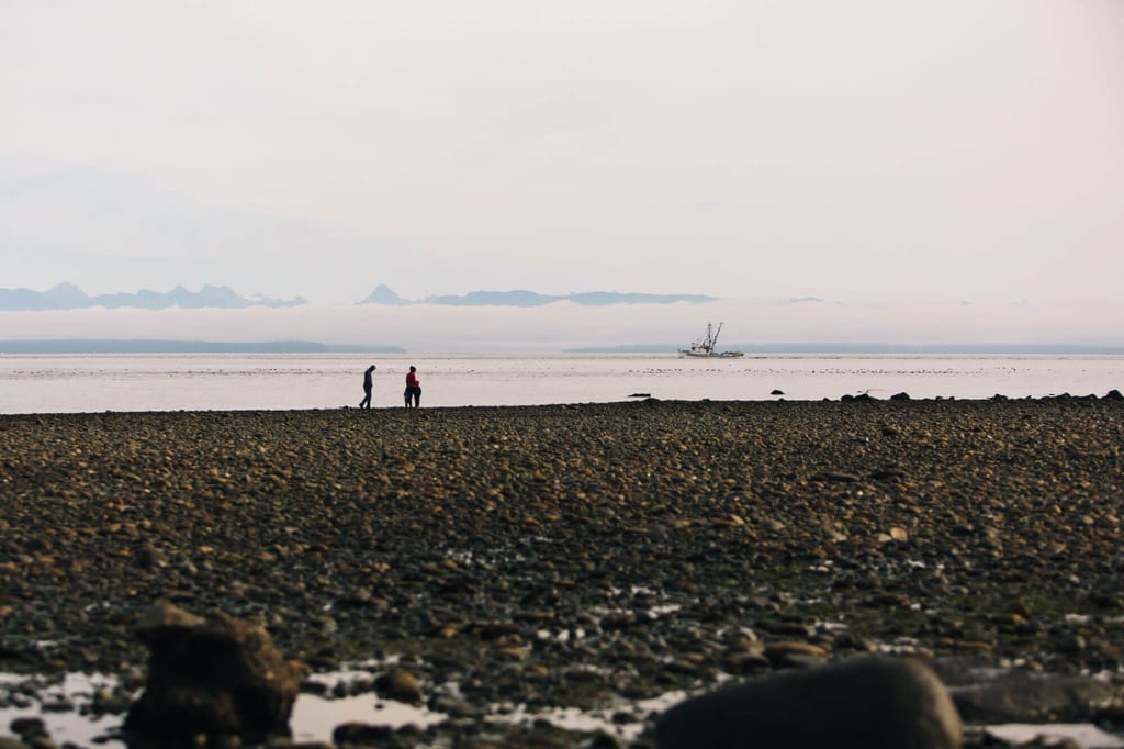 Steiniger Strand mit zwei Personen, entferntes Fischerboot und Bergsilhouette im diffusen Licht