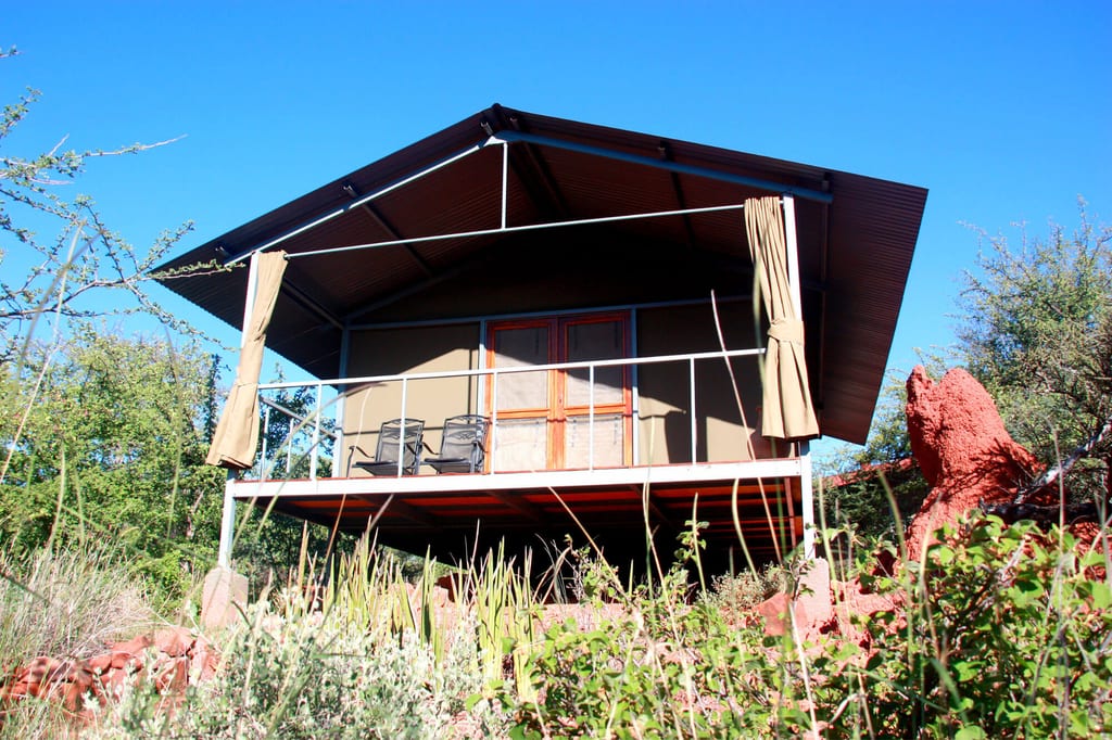 Erhöhtes Safari-Zelt mit Veranda, Stühlen und rotem Termitenhügel vor blauem Himmel