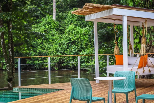 Holzterrasse am Fluss mit Pool, Pavillon und türkisfarbener Sitzgruppe vor dichtem Regenwald
