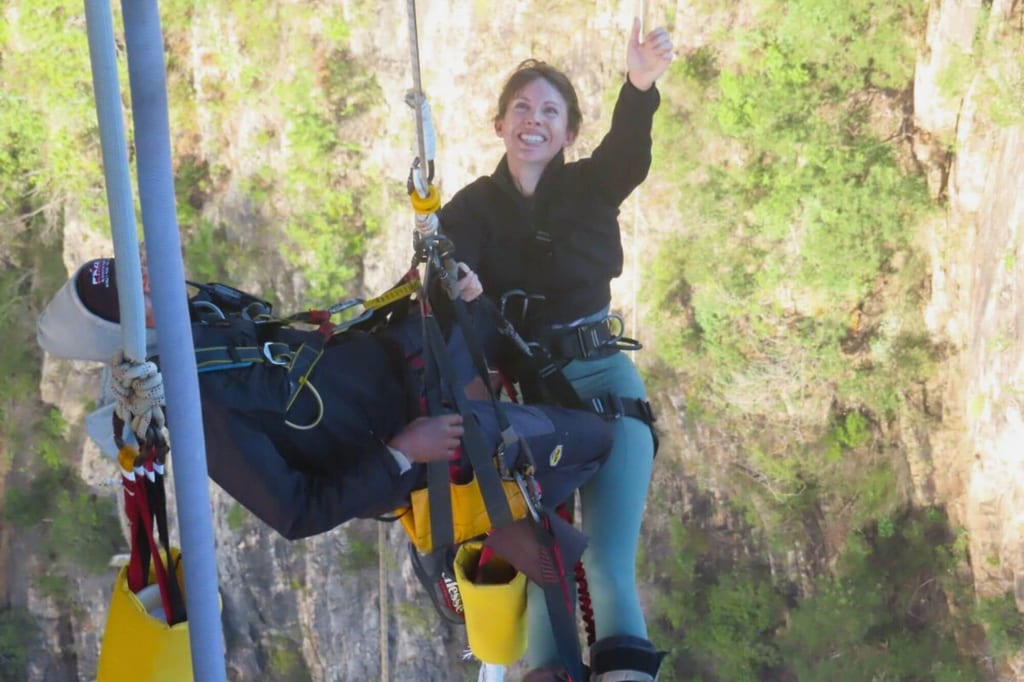 Zwei Personen in Gurten hängen über felsiger Schlucht, eine winkt lächelnd.