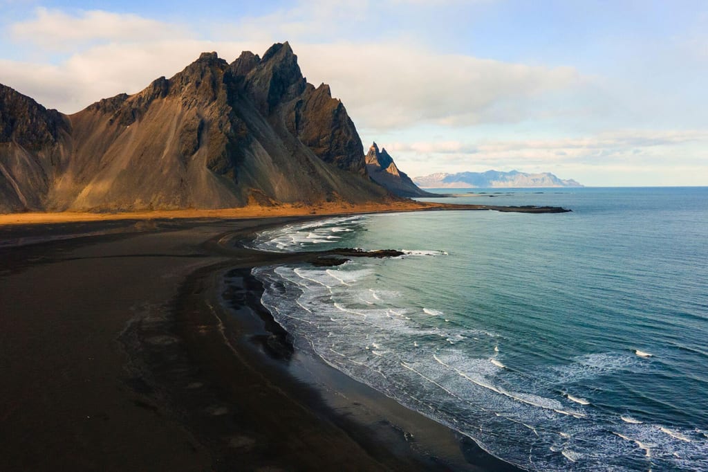 Schroffe Berge über schwarzem Sandstrand und ruhigem Meer im warmen Licht des späten Nachmittags.