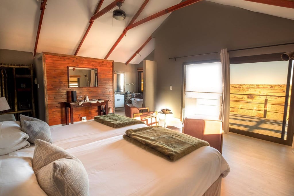 Helles Lodge-Zimmer mit zwei Betten, Holzakzent und offener Tür zur Terrasse mit weiter, trockener Landschaft.