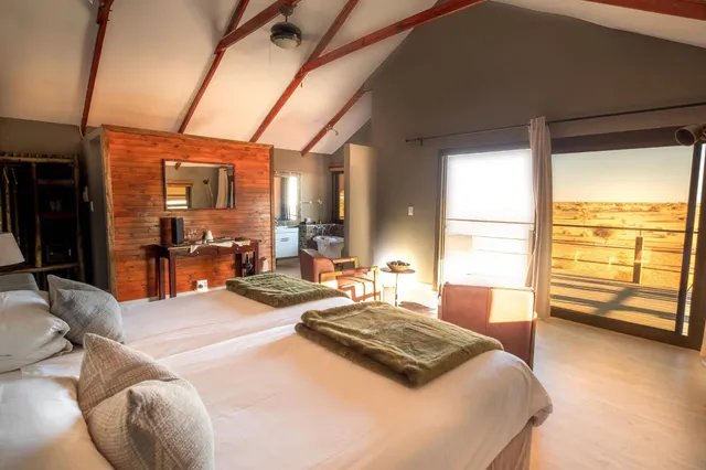 Helles Lodge-Zimmer mit zwei Betten, Holzakzent und offener Tür zur Terrasse mit weiter, trockener Landschaft.
