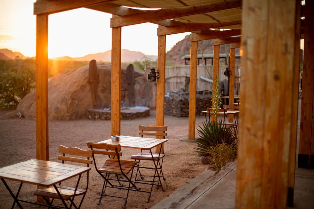Überdachte Holzterrasse mit Tischen und Stühlen in warmem Abendlicht vor felsiger, trockener Landschaft.
