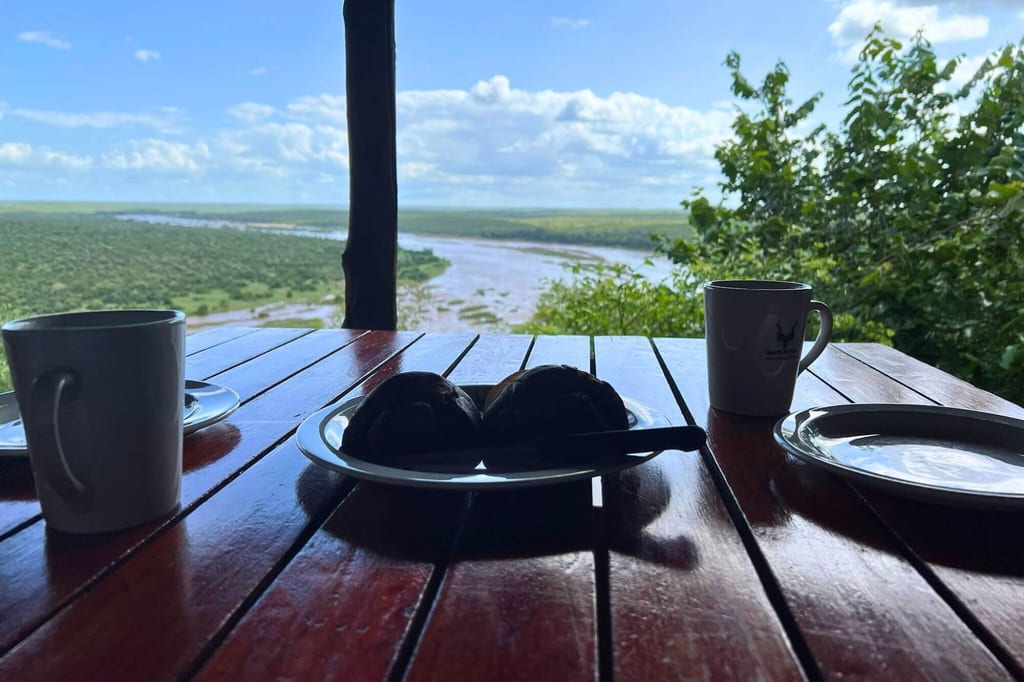 Tisch mit Kaffeetassen und Gebäck, im Hintergrund Fluss und grüne Landschaft unter wolkigem Himmel