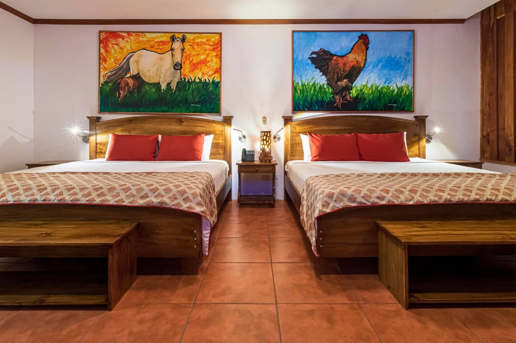 Symmetrisches Hotelzimmer mit zwei Doppelbetten, Holzmöbeln und bunten Tiergemälden über den Betten.