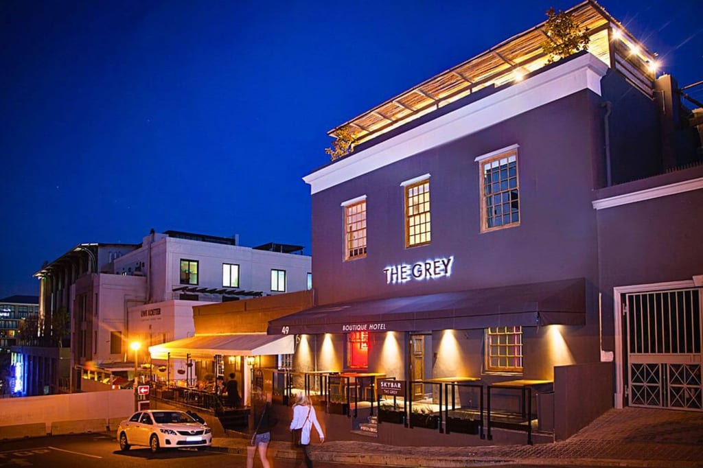Boutique-Hotel mit Leuchtschrift und beleuchteter Terrasse in der blauen Stunde, Fußgänger auf dem Bürgersteig.
