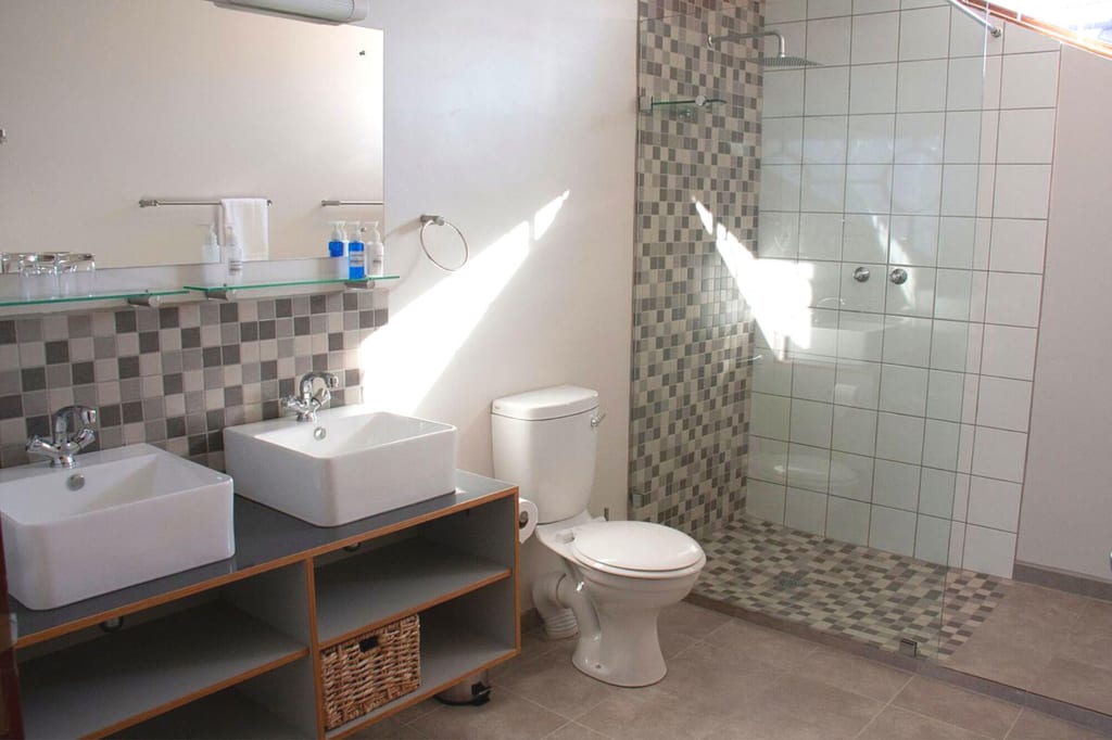 Helles Badezimmer mit zwei Waschbecken, Glasdusche mit Mosaikfliesen und WC, Sonnenlicht fällt ein.