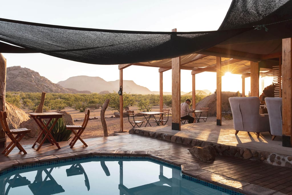Offene Lodge-Terrasse mit Pool, Person an Tisch und warmem Abendlicht über felsiger Landschaft.