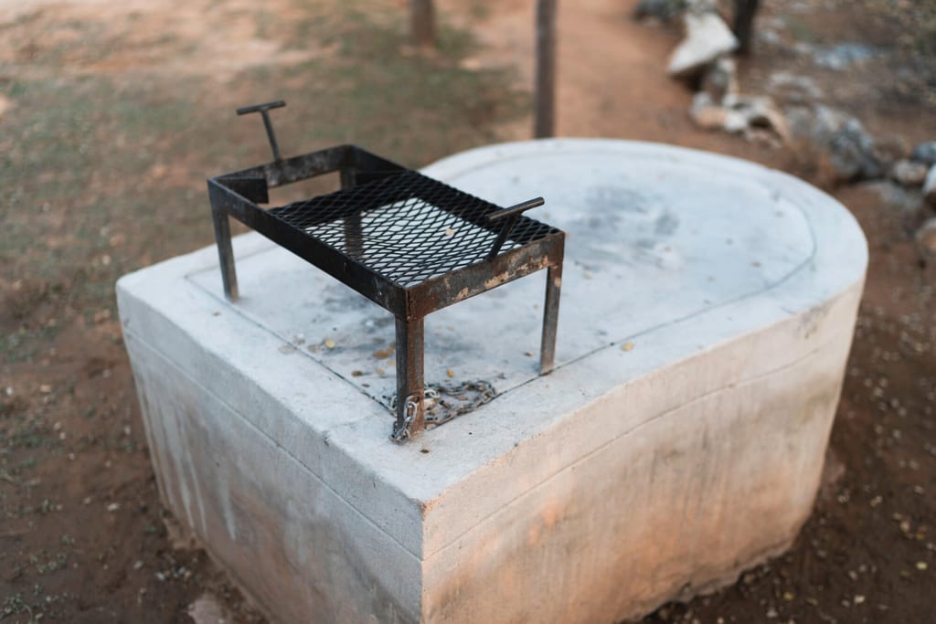 Schwarzes Grillrost auf ovalem Betonsockel im warmen Abendlicht, umgeben von trockenem Boden.