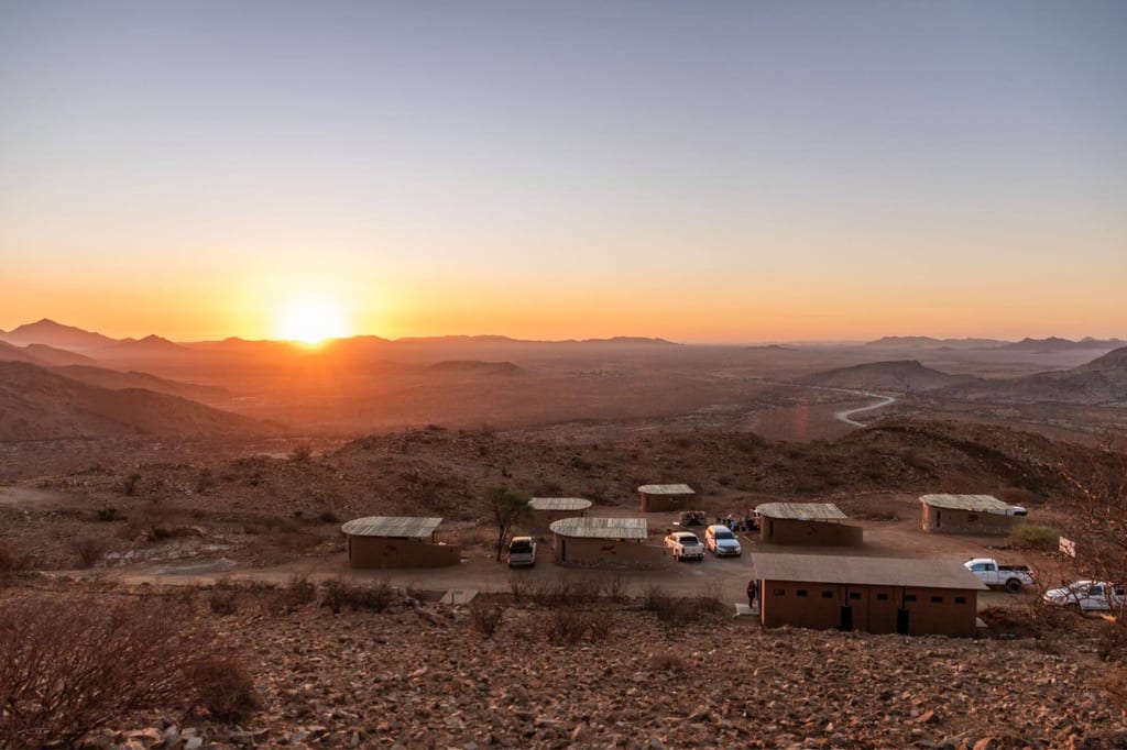 Felsige Halbwüste bei Sonnenaufgang mit kleinem Camp aus Gebäuden und Fahrzeugen, weite Hügel im Hintergrund.