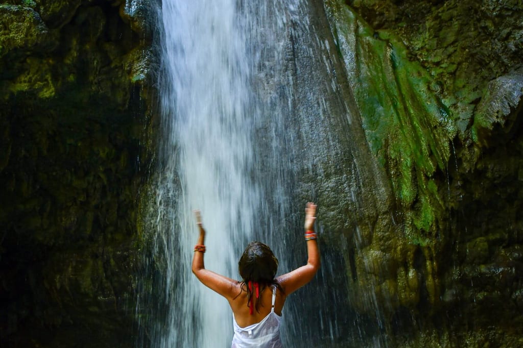 Person mit erhobenen Armen steht unter einem Wasserfall vor moosbewachsenen Felsen.
