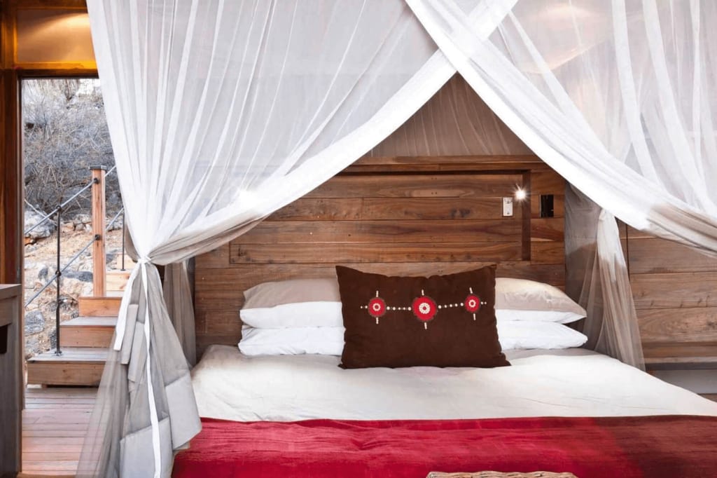 Gemütliches Bett mit Moskitonetz und roter Decke in holzverkleidetem Zelt, offener Eingang zur trockenen Landschaft.