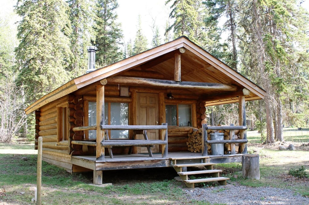 Blockhütte mit Veranda und gestapeltem Brennholz im Nadelwald, Frontansicht bei Tageslicht.