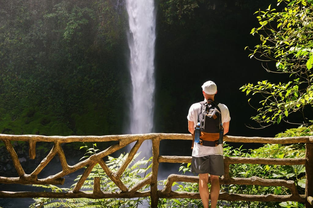Person mit Rucksack steht am Holzgeländer und blickt auf hohen Wasserfall vor grüner Vegetation.