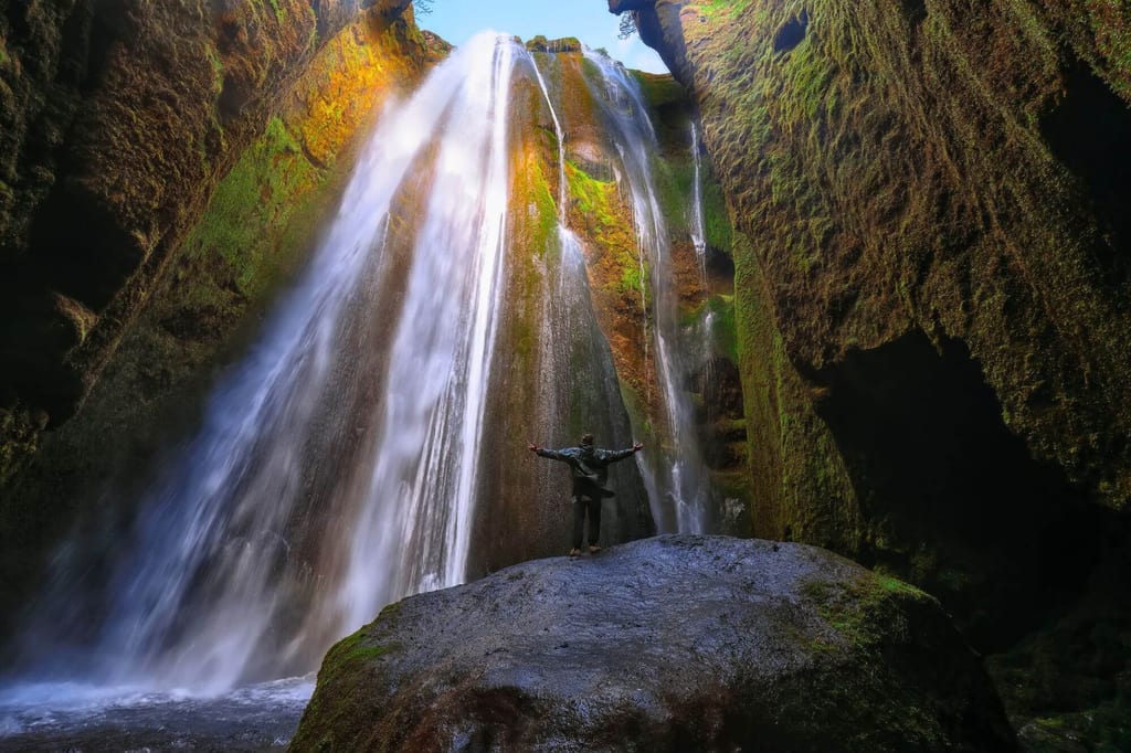 Person mit ausgebreiteten Armen auf Felsen vor moosbedecktem Wasserfall in schmaler Schlucht