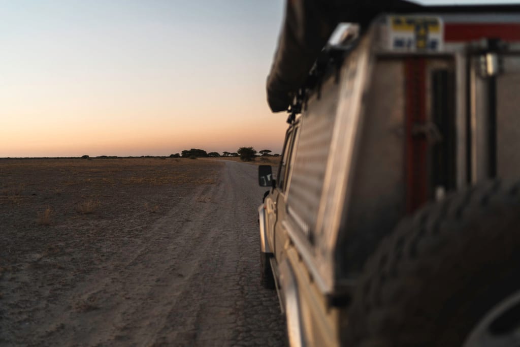 Seitlicher Blick vom Geländewagen über eine sandige Piste in die weite Ebene bei Sonnenuntergang.