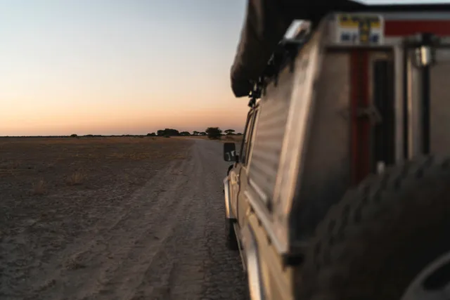 Seitlicher Blick vom Geländewagen über eine sandige Piste in die weite Ebene bei Sonnenuntergang.