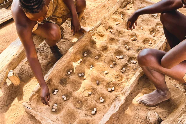 Zwei Personen spielen auf einem Muldenbrett mit kleinen Steinen auf sandigem Boden in warmer Mittagsbeleuchtung.