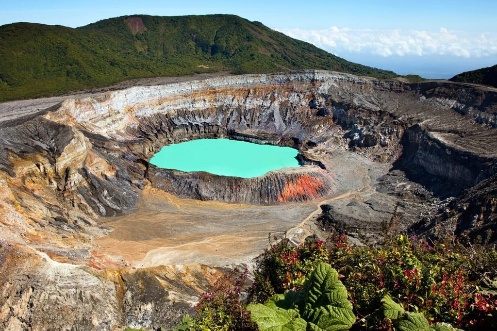 Vulkankrater mit türkisfarbenem Kratersee und Vegetation im Vordergrund bei hellem Tageslicht