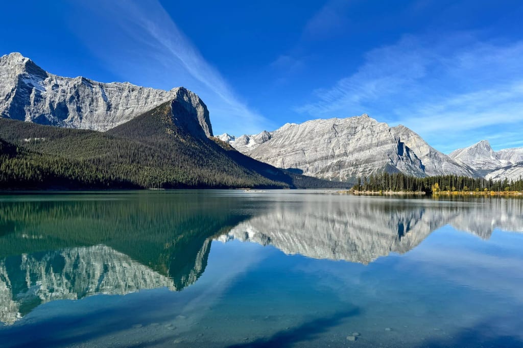 Ruhiger Gebirgssee spiegelt schroffe Berge und Nadelwald unter klarem blauem Himmel.