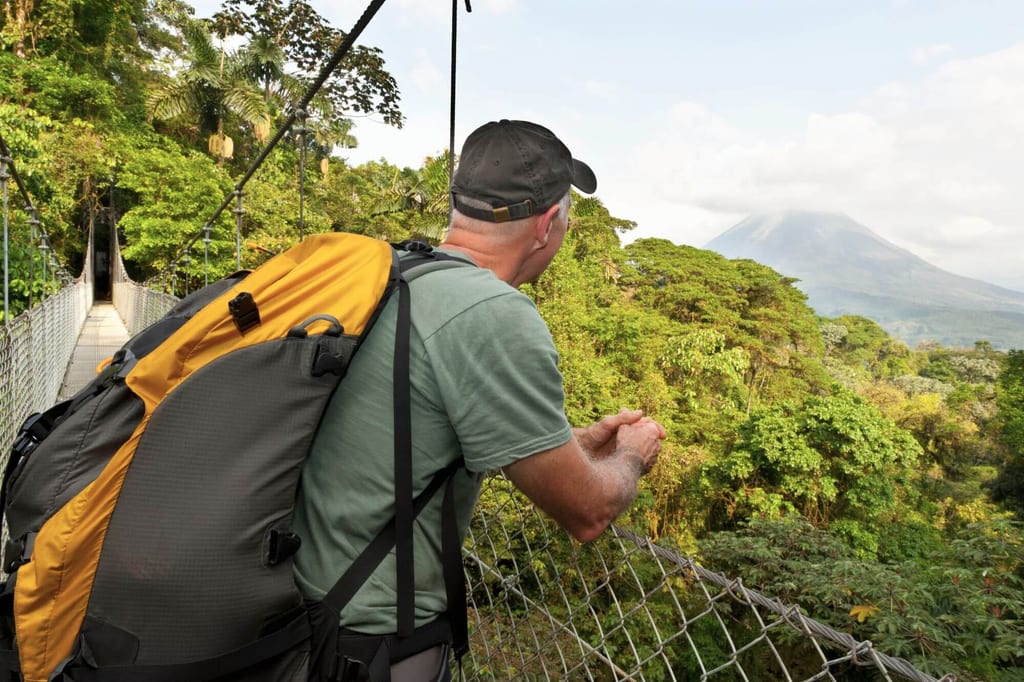 Wanderer mit großem Rucksack auf Hängebrücke blickt über Regenwald zu einem fernen Vulkan
