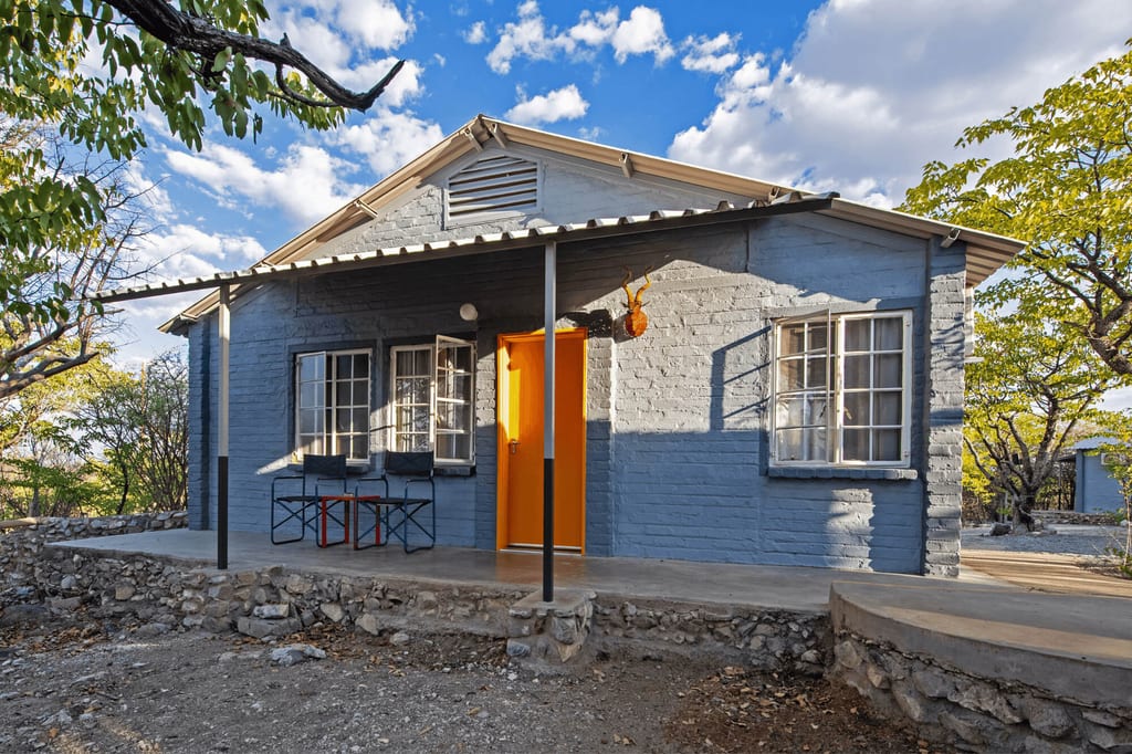 Blaues Steinhaus mit orangefarbener Tür auf steinigem Grund, Veranda und Nachmittagslicht