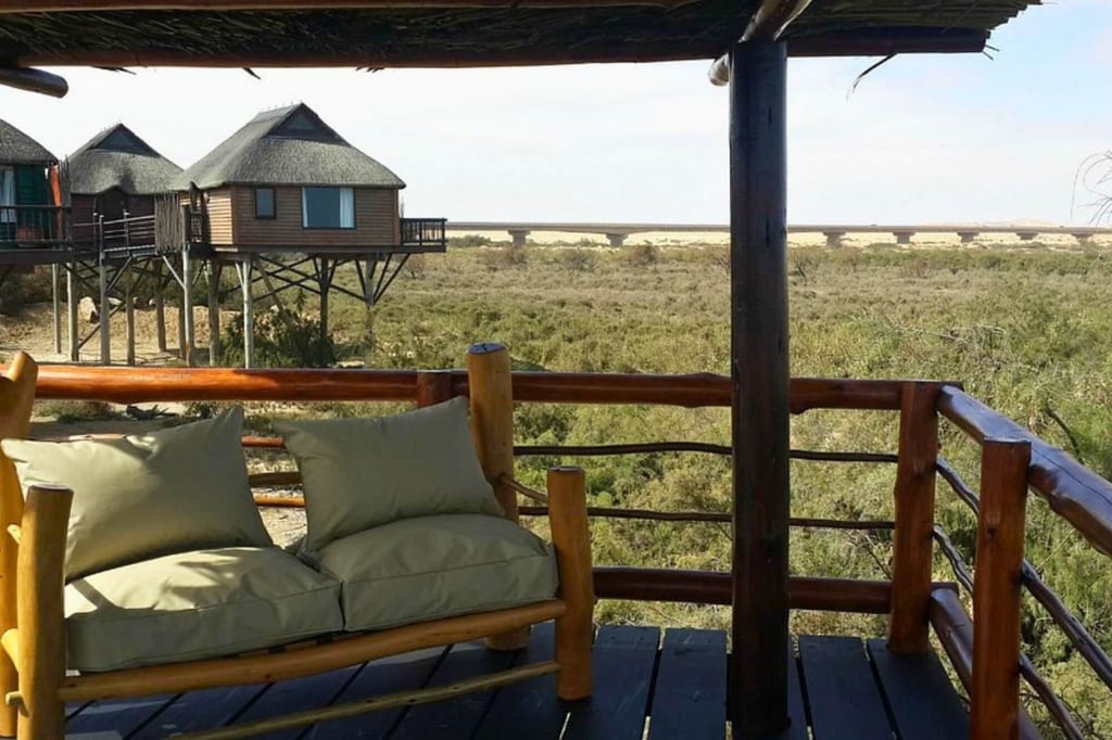 Veranda mit Holzbänkchen und Blick auf strohgedeckte Stelzenhütten und buschige Landschaft