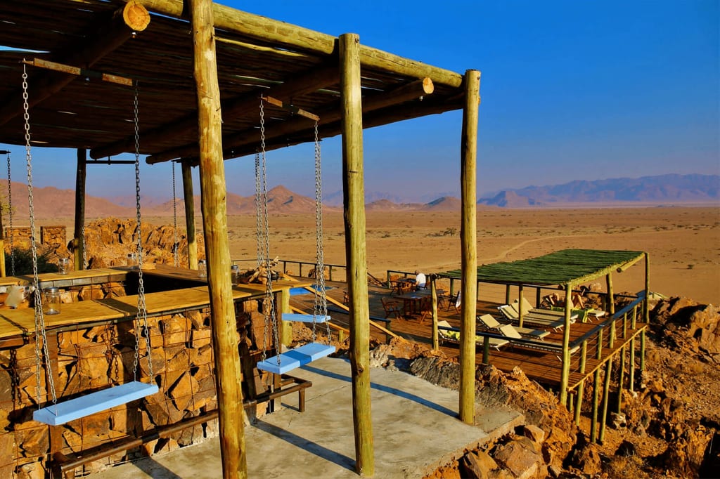 Holzterrasse mit Schaukeln und Liegen an Felsrand über weiter Wüstenebene und Bergen