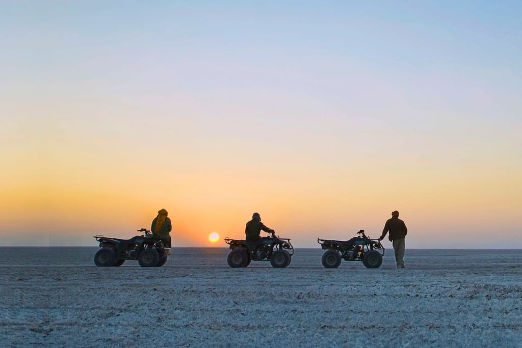 Drei Personen mit Quads als Silhouetten auf weiter Fläche bei Sonnenuntergang
