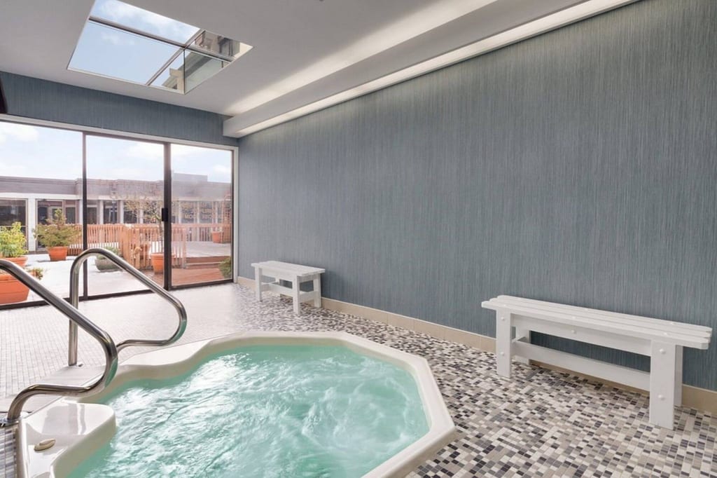 Innenraum mit sprudelndem Whirlpool, Mosaikboden, weißen Bänken und Terrasse im Tageslicht.