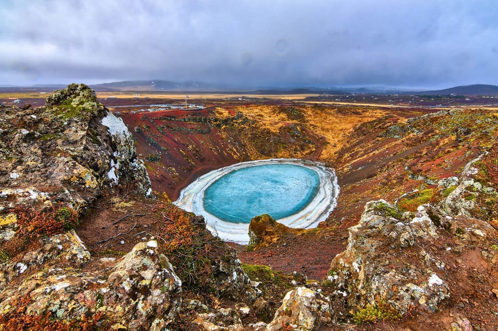 Blick von einem felsigen Kraterrand auf einen kreisrunden, teilweise gefrorenen Kratersee mit türkisblauem Wasser.