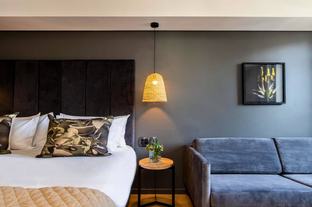 Modernes Hotelzimmer mit Bett, grauem Sofa, Hängeleuchte und botanischem Bild an grauer Wand