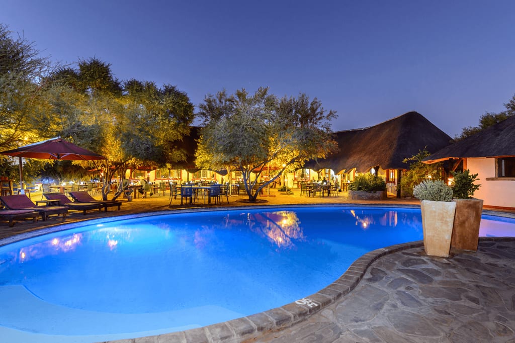 Beleuchteter Lodge-Pool bei Dämmerung mit thatchgedeckten Gebäuden und Sitzgruppen
