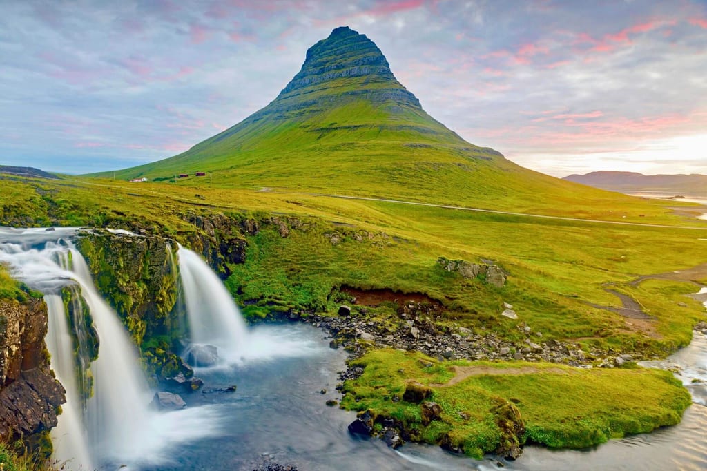 Konischer Berg über grüner Hügellandschaft mit Wasserfall im Vordergrund bei Abendlicht.