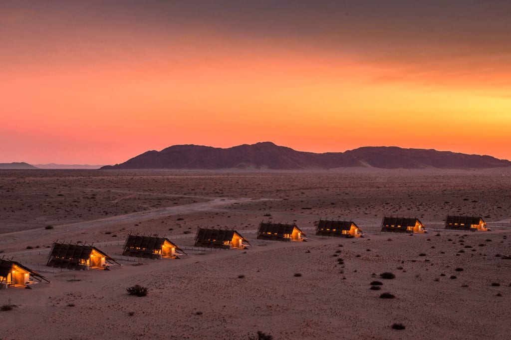 Vogelperspektive auf ein beleuchtetes Wüstencamp bei orange‑rosa Sonnenuntergang vor niedriger Bergsilhouette.