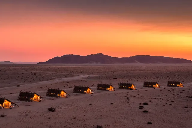 Vogelperspektive auf ein beleuchtetes Wüstencamp bei orange‑rosa Sonnenuntergang vor niedriger Bergsilhouette.
